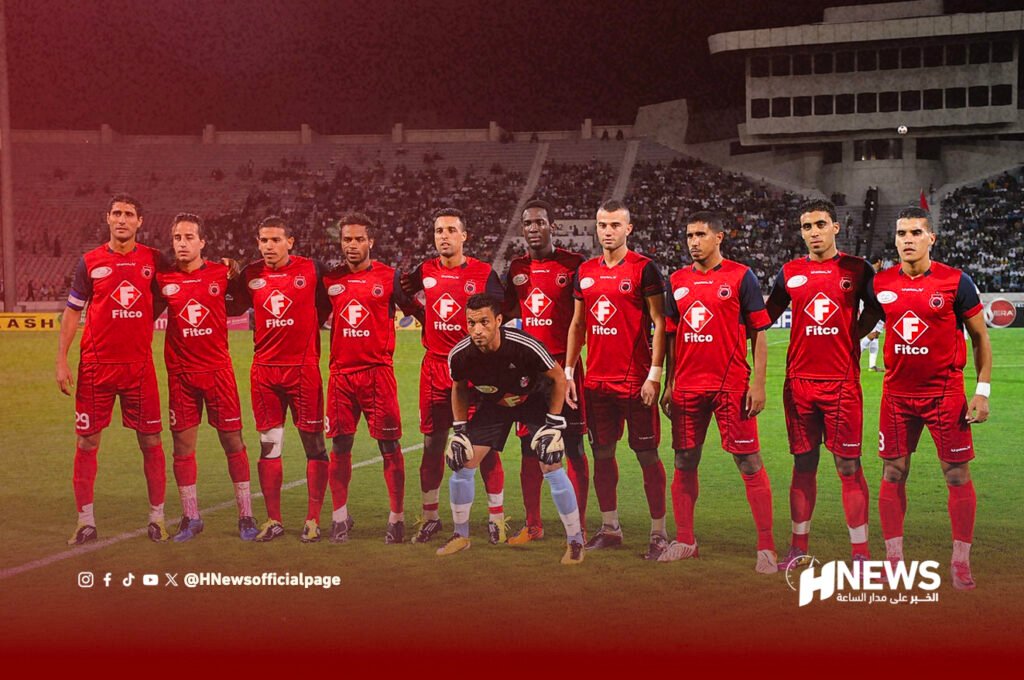 Soccer team lineup posing on the field with goalkeeper crouching center, all players in red uniforms with Fitco sponsor, stadium stands behind them.