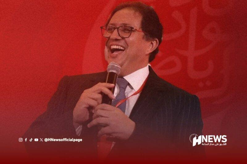 Smiling man in a dark suit and glasses speaks into a microphone on stage, with a red backdrop and HNews logo behind him.