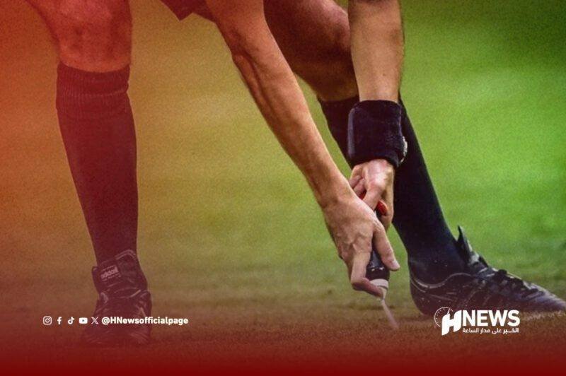 Close-up of a rugby player adjusting a kicking tee on a grassy field; hands on the ball, wearing red and black socks, HNews logo bottom right.