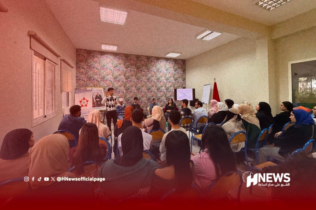 Speaker addressing a seated audience in a conference room with banners and a screen at the front, and the HNews logo in the bottom-right corner.