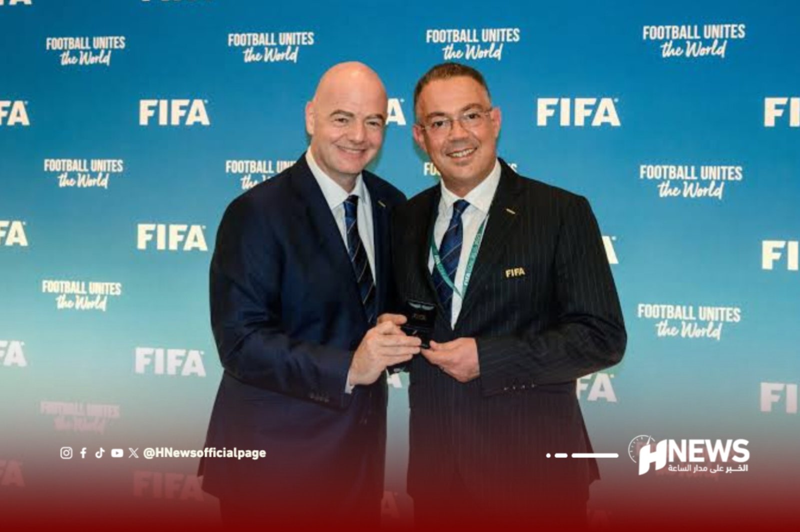 Two men in suits pose with a trophy at a FIFA event against a blue backdrop filled with FIFA logos and the slogan 'Football Unites the World.'