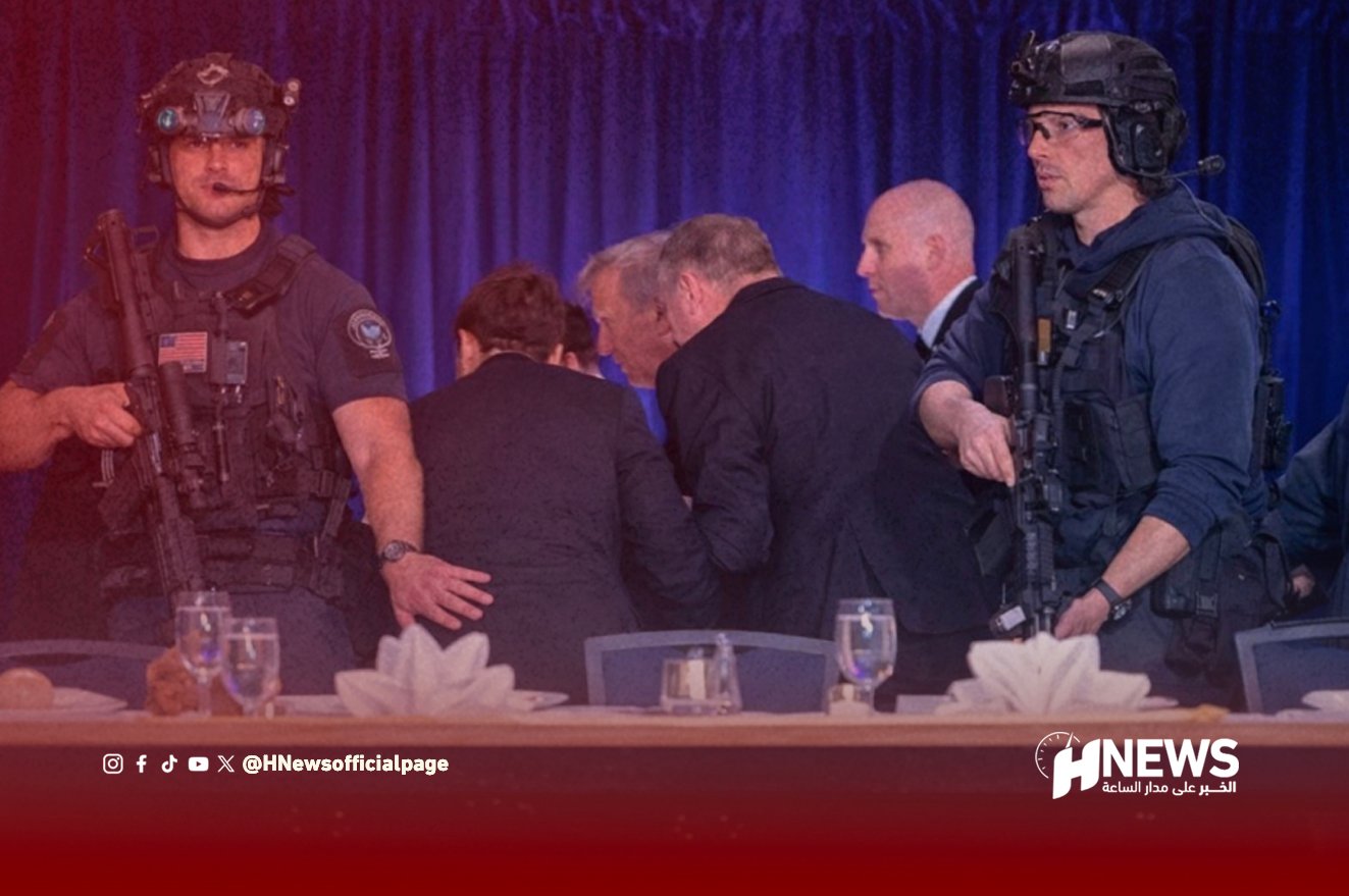 Two armed security officers in tactical gear stand beside a group of suited men at a table on stage.