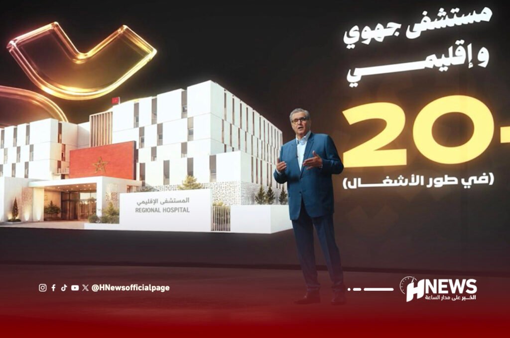 Presenter in a blue suit stands on stage beside a large rendering of a modern regional hospital, with Arabic text and the HNews logo in the background.
