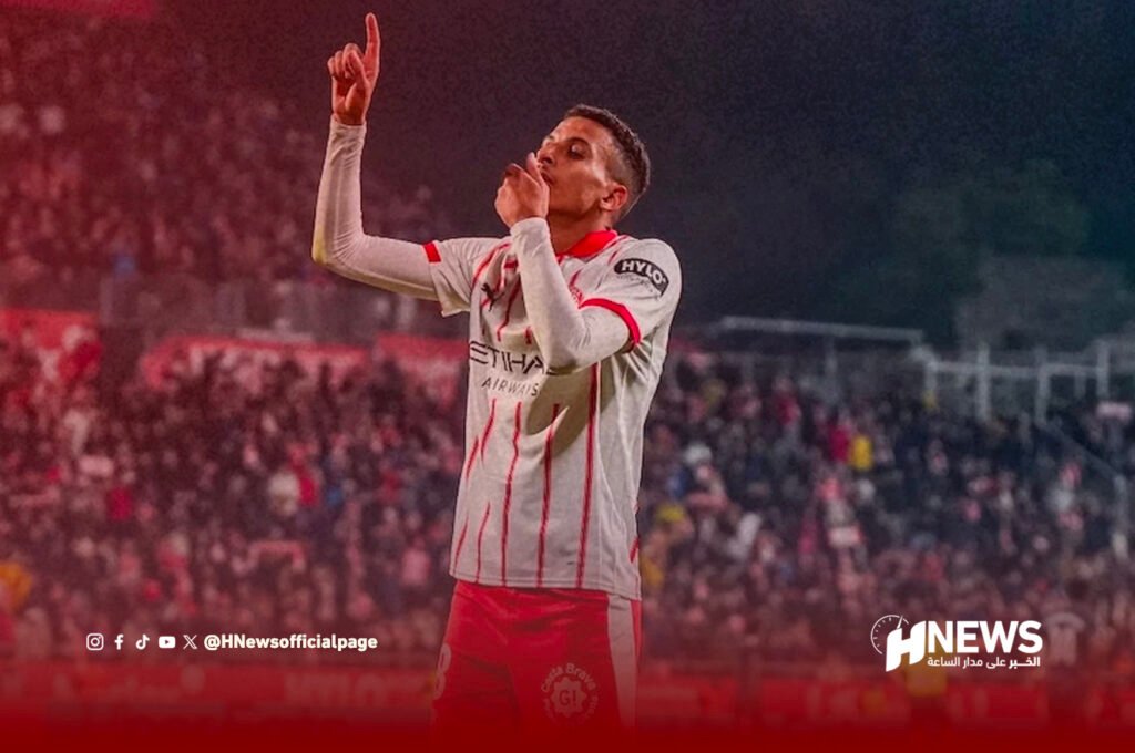 أوناحي | H-NEWS آش نيوز Soccer player in a white and red kit celebrates a goal, raising one finger and touching his lips as fans cheer in the background.