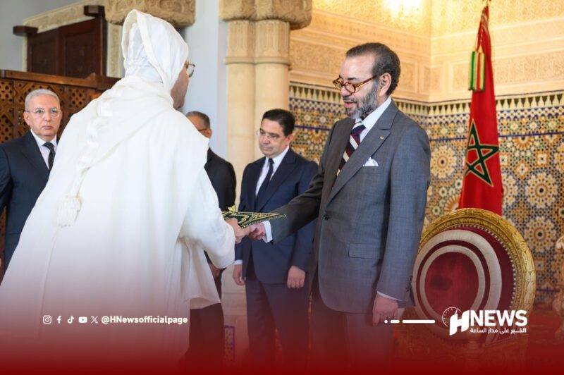 A man in a gray suit hands a ceremonial item to a person in a white traditional robe during a formal ceremony, with a Moroccan flag and ornate decor in the background.