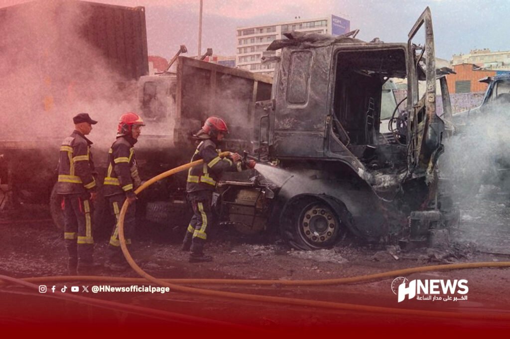 Firefighters in protective gear spray water on a burned, smoking truck on a city street.