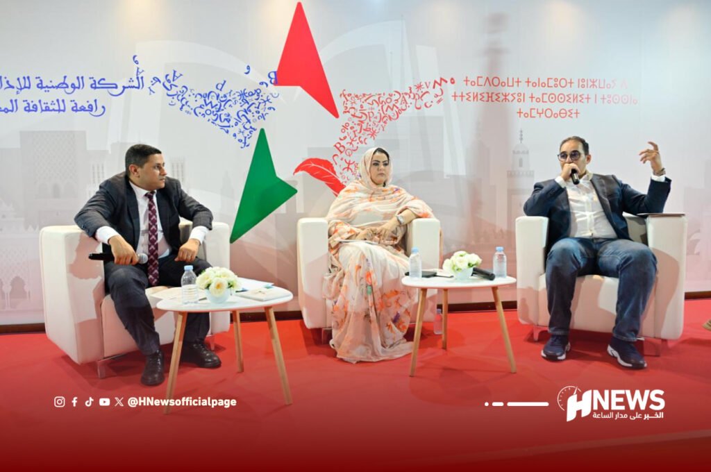 Panel discussion with three speakers seated in white chairs on a red carpet, microphones in hand, backdrop with abstract shapes and Arabic text behind them.