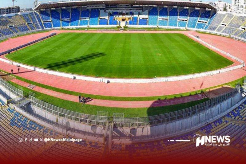 Empty outdoor football stadium with a green pitch, red running track around it, and blue spectator stands.