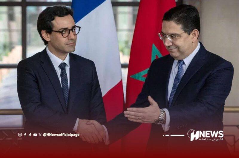 وزيرا-الخارجية-المغربي-والفرنسي-scaled | H-NEWS آش نيوز Two men in dark suits shake hands in a formal setting with France and Morocco flags behind them.