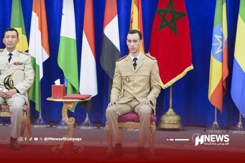 Two uniformed men sit on ornate chairs at an official ceremony, with multiple national flags behind them and a red carpet scene behind.