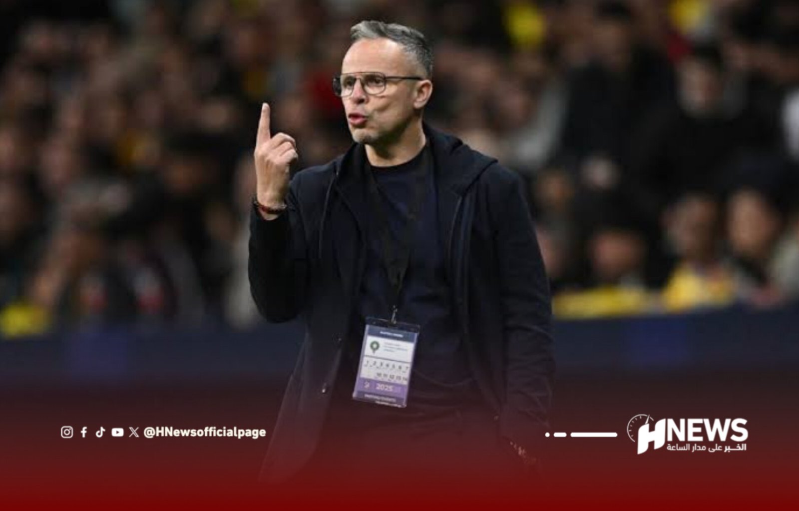 Man in dark jacket with a lanyard badge on a blurred crowd background, gesturing with his index finger raised as if making a point at an event site.