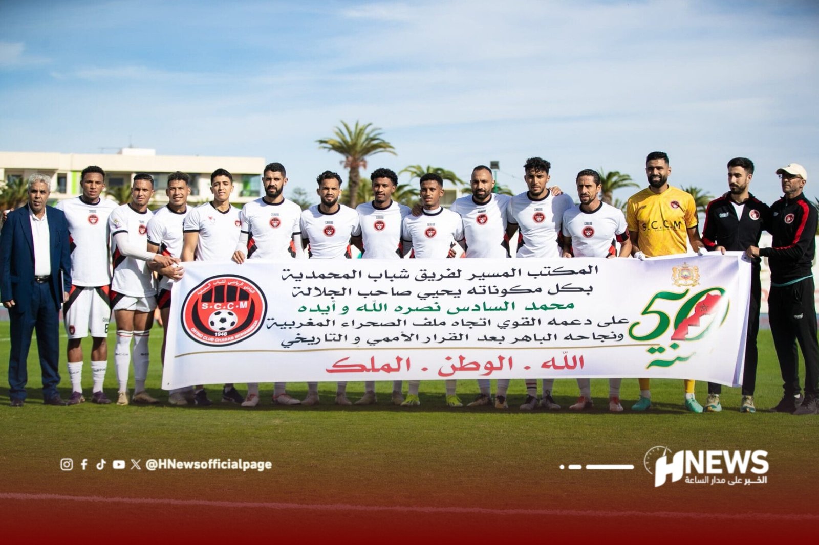 Soccer players in white uniforms posing with a banner on a field, group photo with coaches and staff against a blue sky.