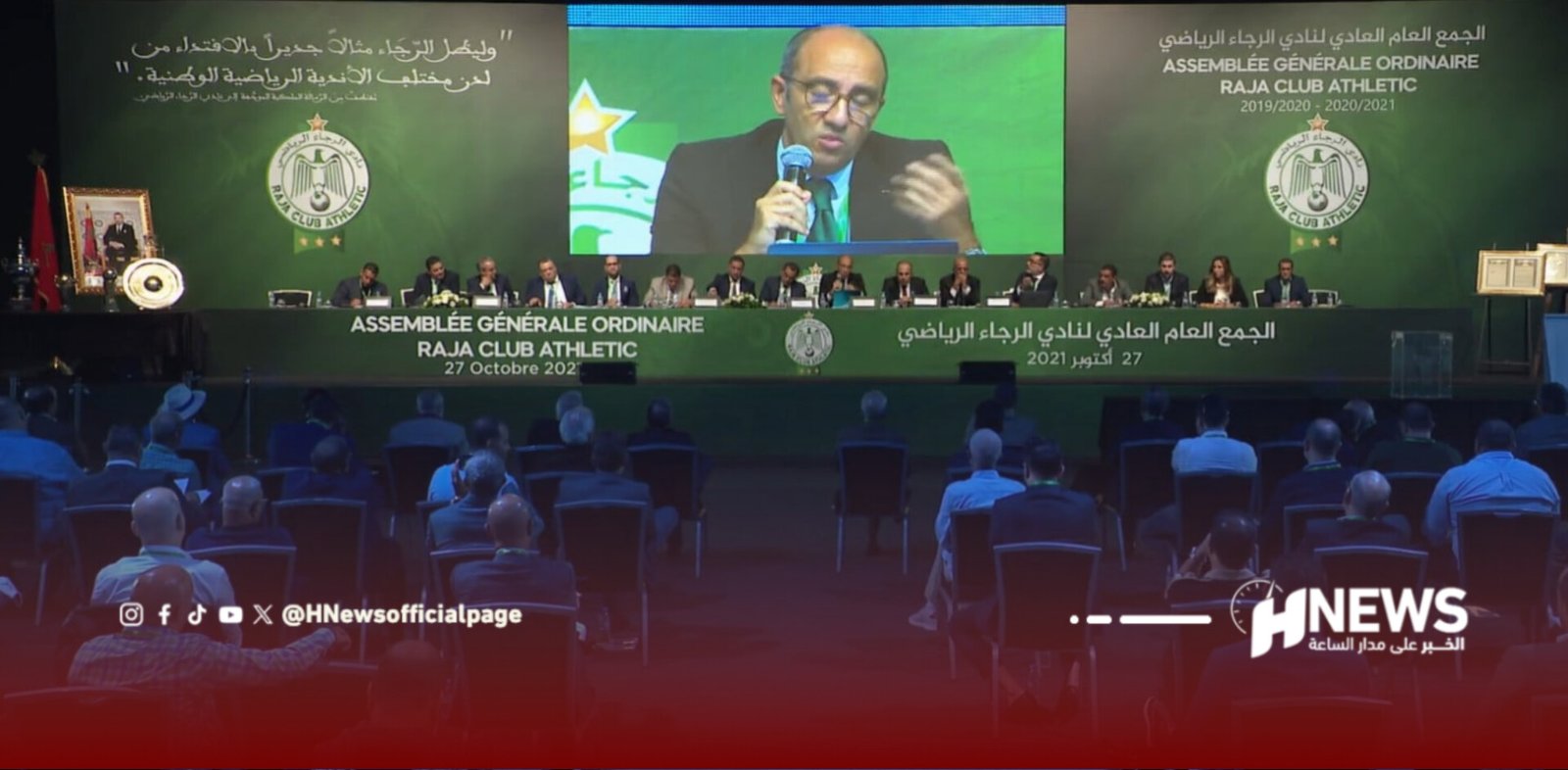 Panel of officials seated at a long table on stage with a large screen showing a speaking man and Raja Club Athletic banners in Arabic/French behind him.
