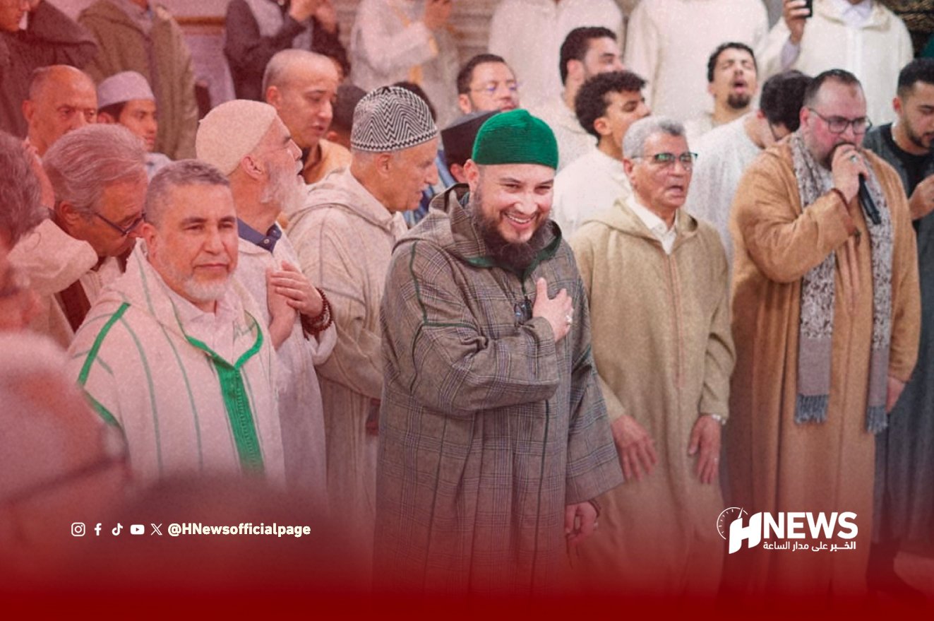 Smiling man in a green cap places a hand on his chest among a crowd of men in traditional robes.