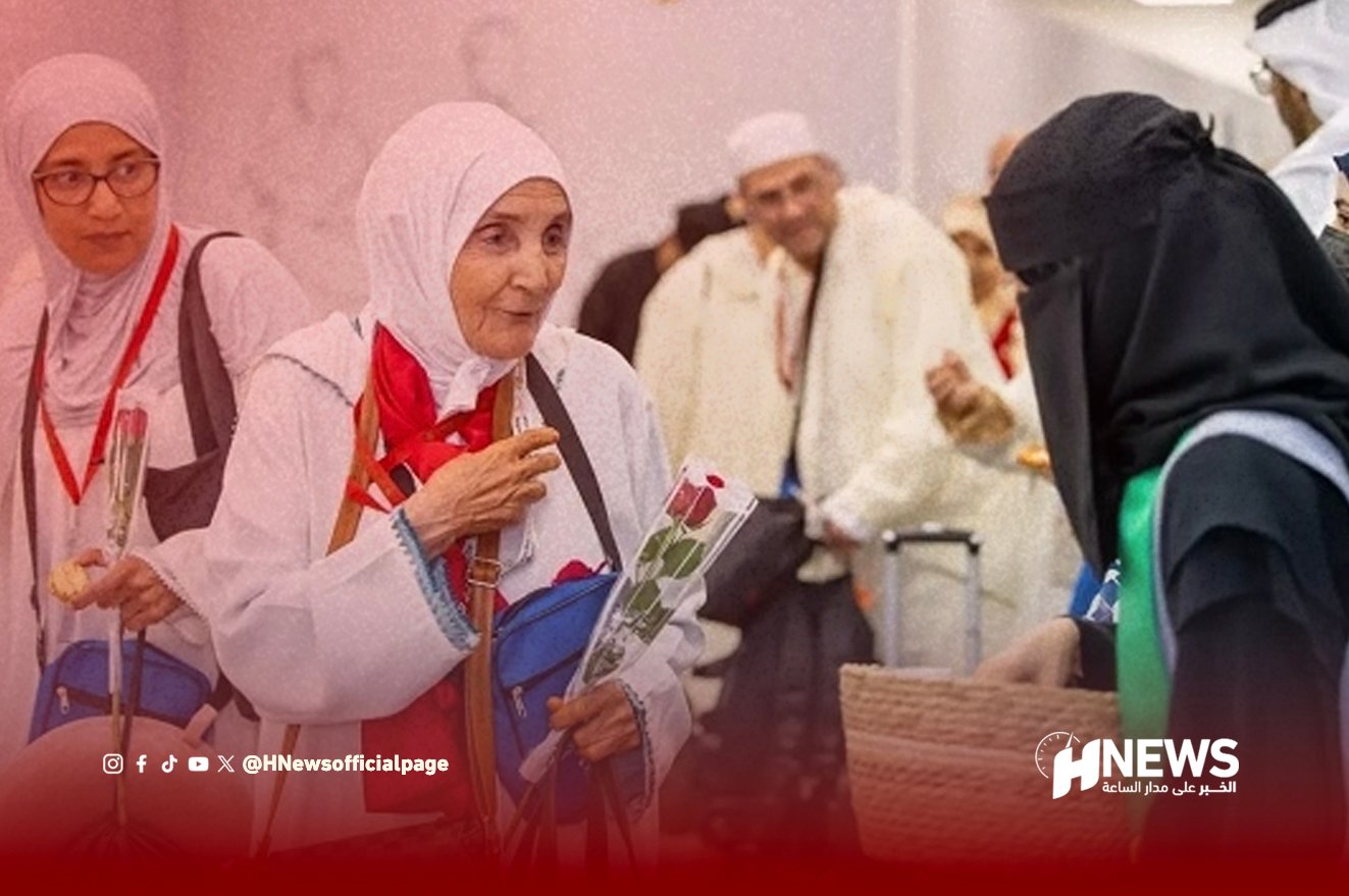 Elderly woman in a white hijab holding flowers chats with a staff member in a black hijab at a service counter, with onlookers in the background.