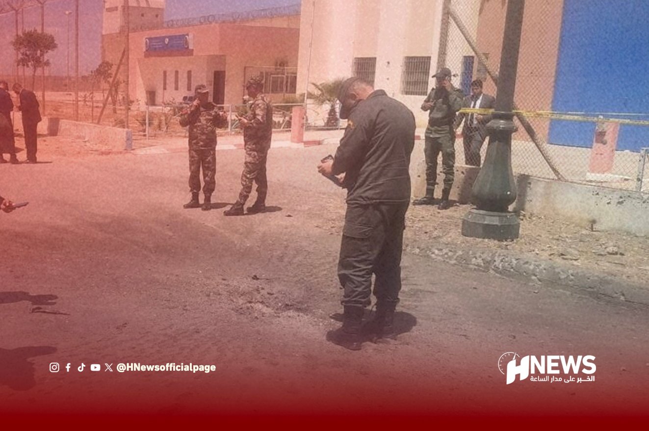 Law enforcement officers stand behind yellow tape at a cordoned street with rubble and a large lamp post; news watermark visible.