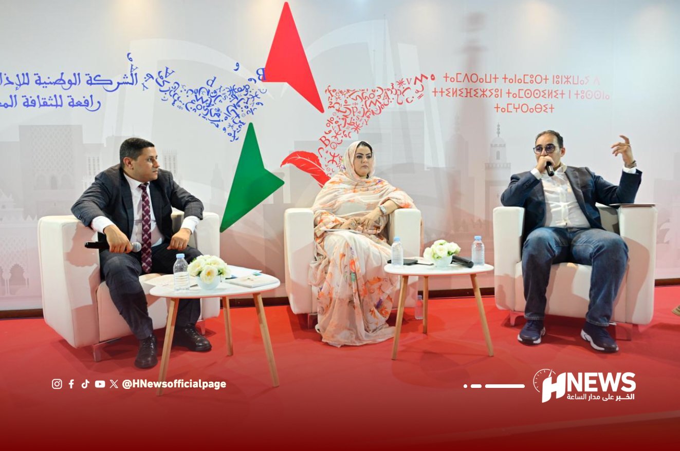 Panel discussion with three speakers seated in white chairs on a red carpet, microphones in hand, backdrop with abstract shapes and Arabic text behind them.