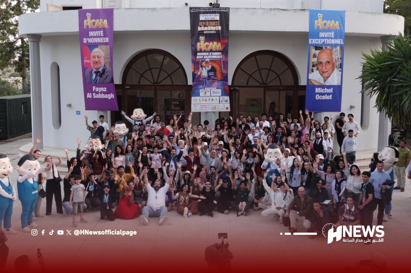 Large group of people posing for a group photo in front of a modern building with three event banners and costumed mascots.",