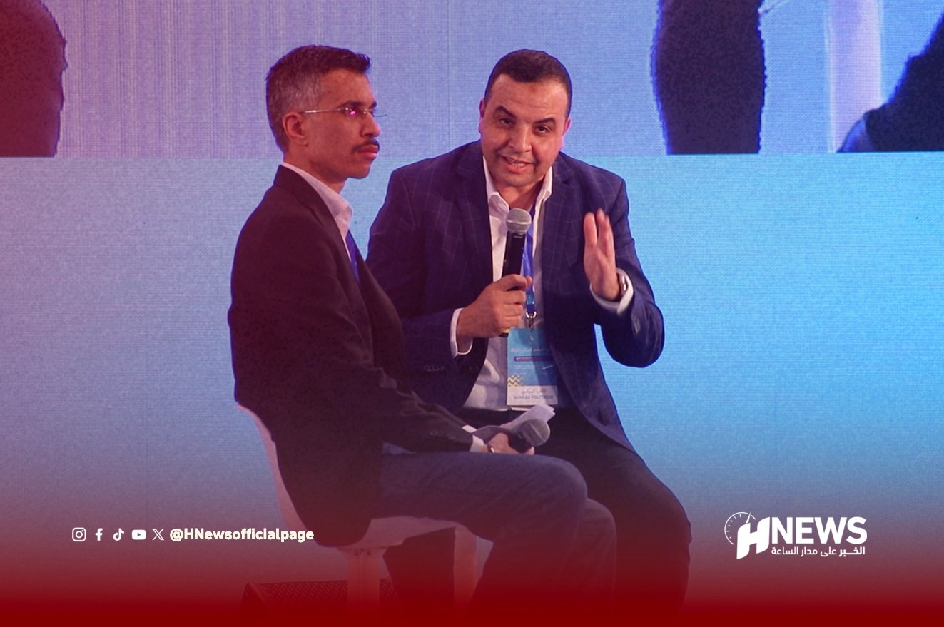 Two men in suits seated on a stage panel; one is speaking into a microphone while the other listens, with a blue backdrop and HNews watermark in the corner.