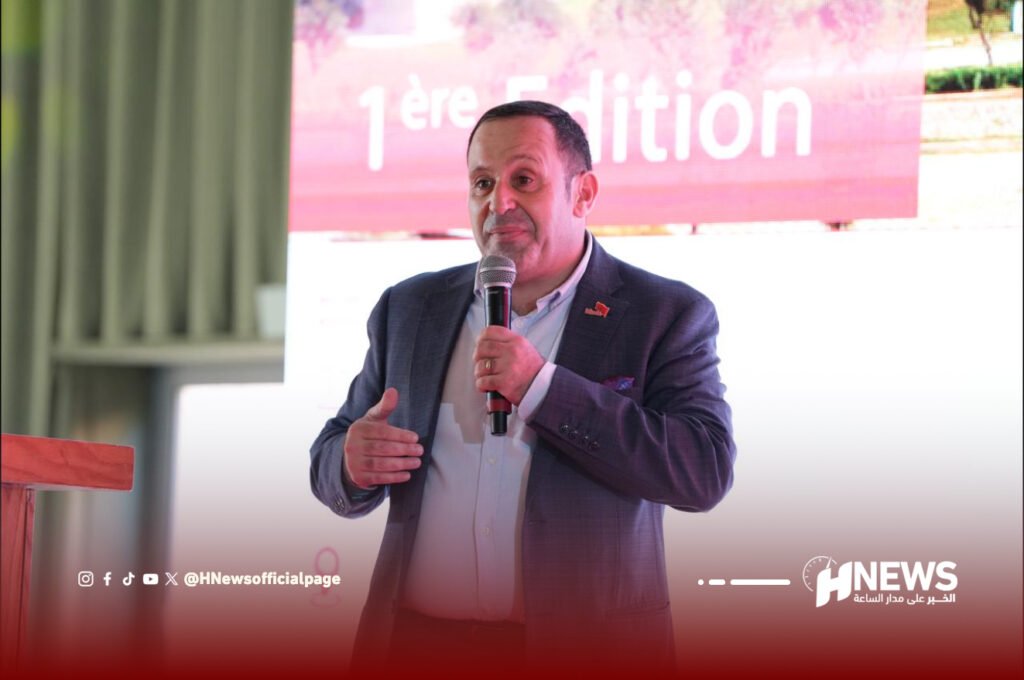 أدوية الحكمة | H-NEWS آش نيوز Man in a suit speaking into a handheld microphone on stage, with a pink banner behind him that reads '1ère édition' and a watermark 'HNews' at the bottom right.