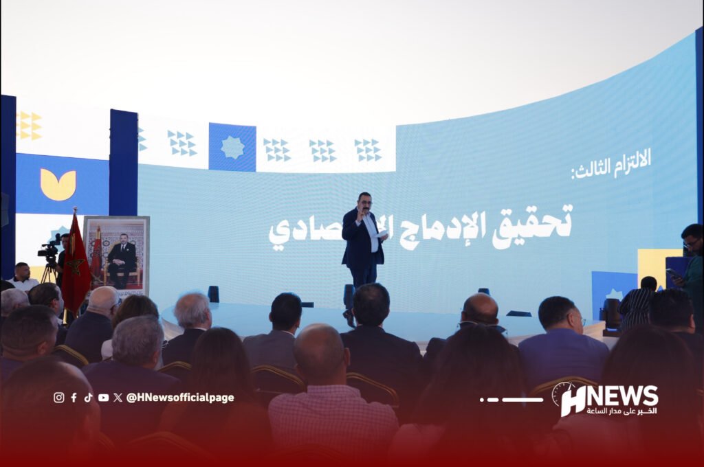 الشافقي | H-NEWS آش نيوز Speaker on a blue stage delivering a talk to a seated audience, with a large Arabic banner behind reading during the third commitment and HNews logo at the front-right.