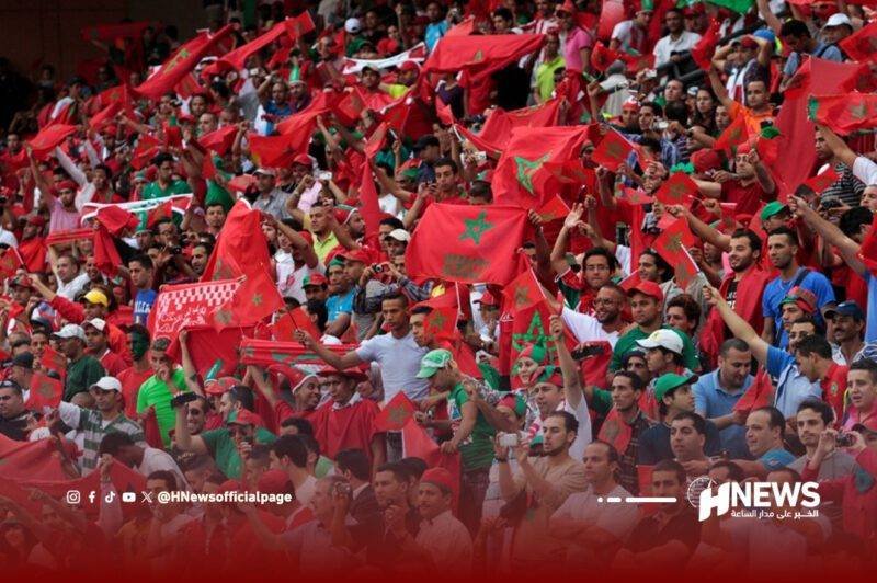 جمهور-المنتخب-النغربي-scaled | H-NEWS آش نيوز Large crowd of fans in red, waving flags with a green star at a stadium, cheering and filling the stands.