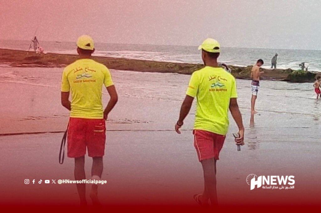 سباح منقذ | H-NEWS آش نيوز Two lifeguards in neon yellow shirts and red shorts walk along a sandy beach at the shoreline.