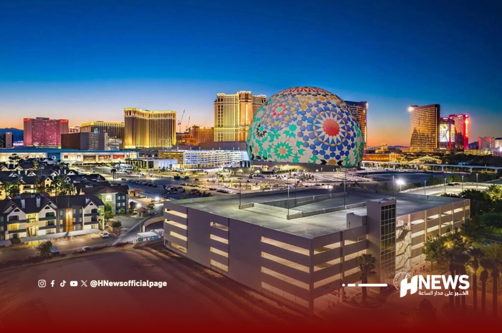شاشة المغرب لاس فيغاس | H-NEWS آش نيوز Aerial night view of a city with a large colorful mosaic dome among high-rise buildings and a parking garage in the foreground, with social media watermark in the corner.