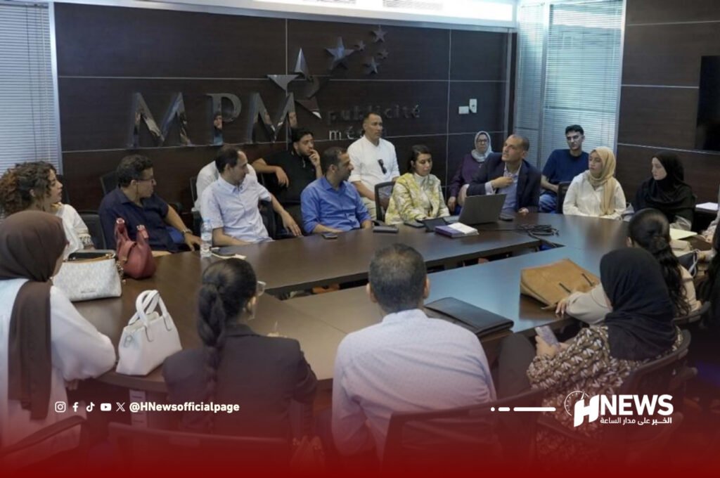 Group of adults around a large conference table in a boardroom, some with laptops, discussing a presentation; a logo wall behind them reads 'MPM'.