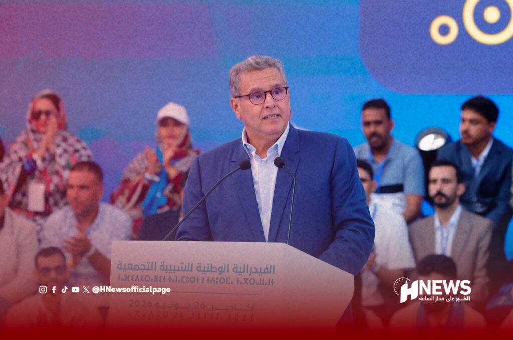 Man in a blue suit delivering a speech at a podium with two microphones, audience and media in the blurred background, HNews logo visible bottom right.