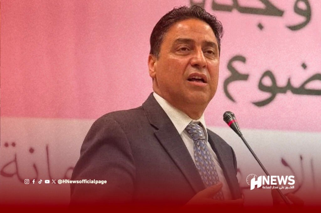 عمر حجيرة | H-NEWS آش نيوز Man in a dark suit and patterned tie speaks at a microphone on a stage with a pink backdrop and a news outlet watermark in the corner.