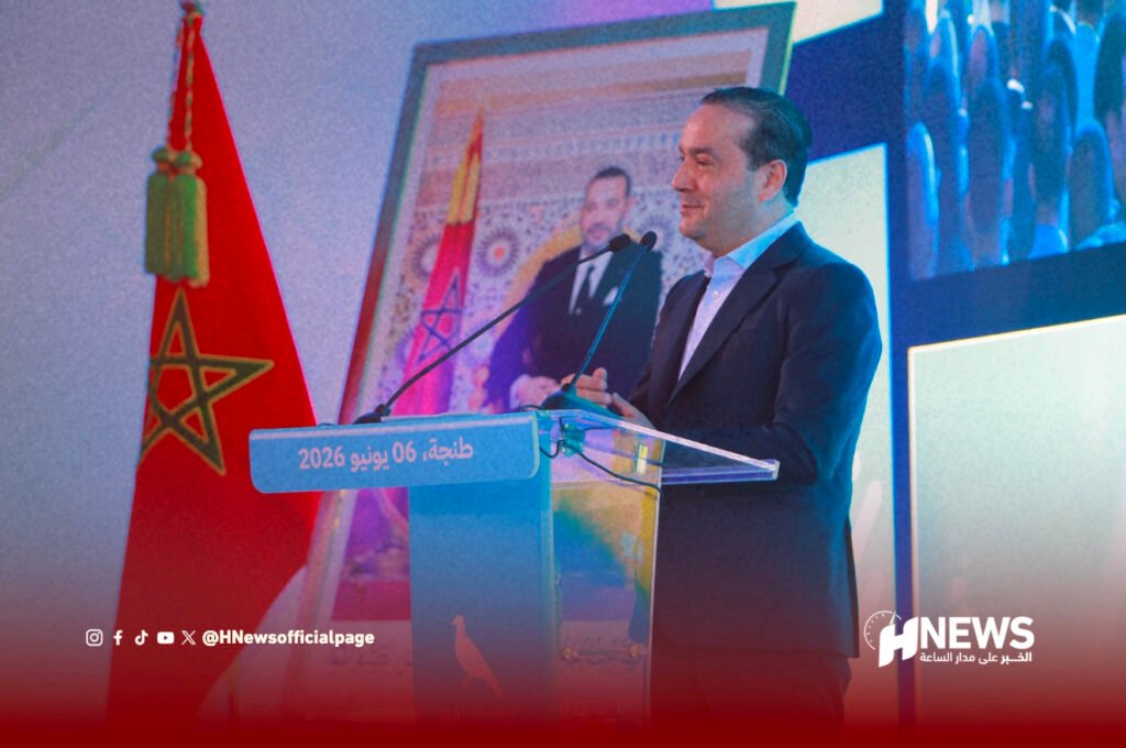 محمد شوكي | H-NEWS آش نيوز Man in a suit speaks at a clear podium during a formal event; Moroccan flag and a framed portrait are in the background (Tangier, 06 June 2026).