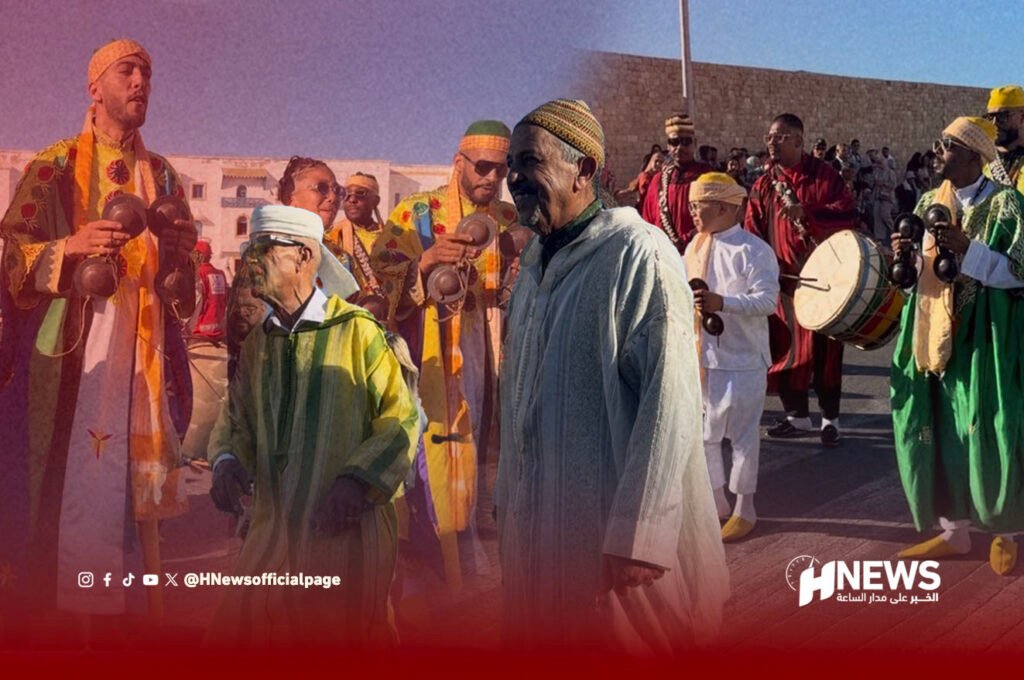 Group of men in colorful traditional robes playing percussion at an outdoor cultural festival, with a drummer and crowd nearby and a red gradient overlay at the bottom (HNEWS logo).