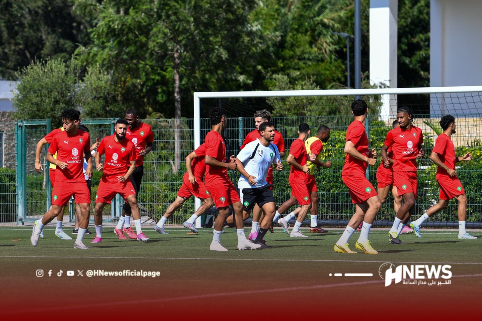 Soccer players in red jerseys run on a practice field near a goal, with a green fence and trees in the background.