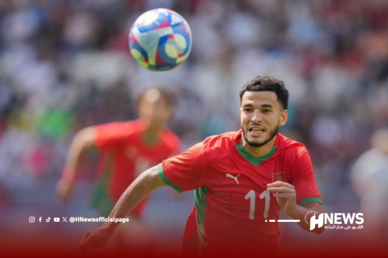 Soccer player in a red jersey (number 11) runs toward a colorful ball in midair during a match, blurred crowd in the background.