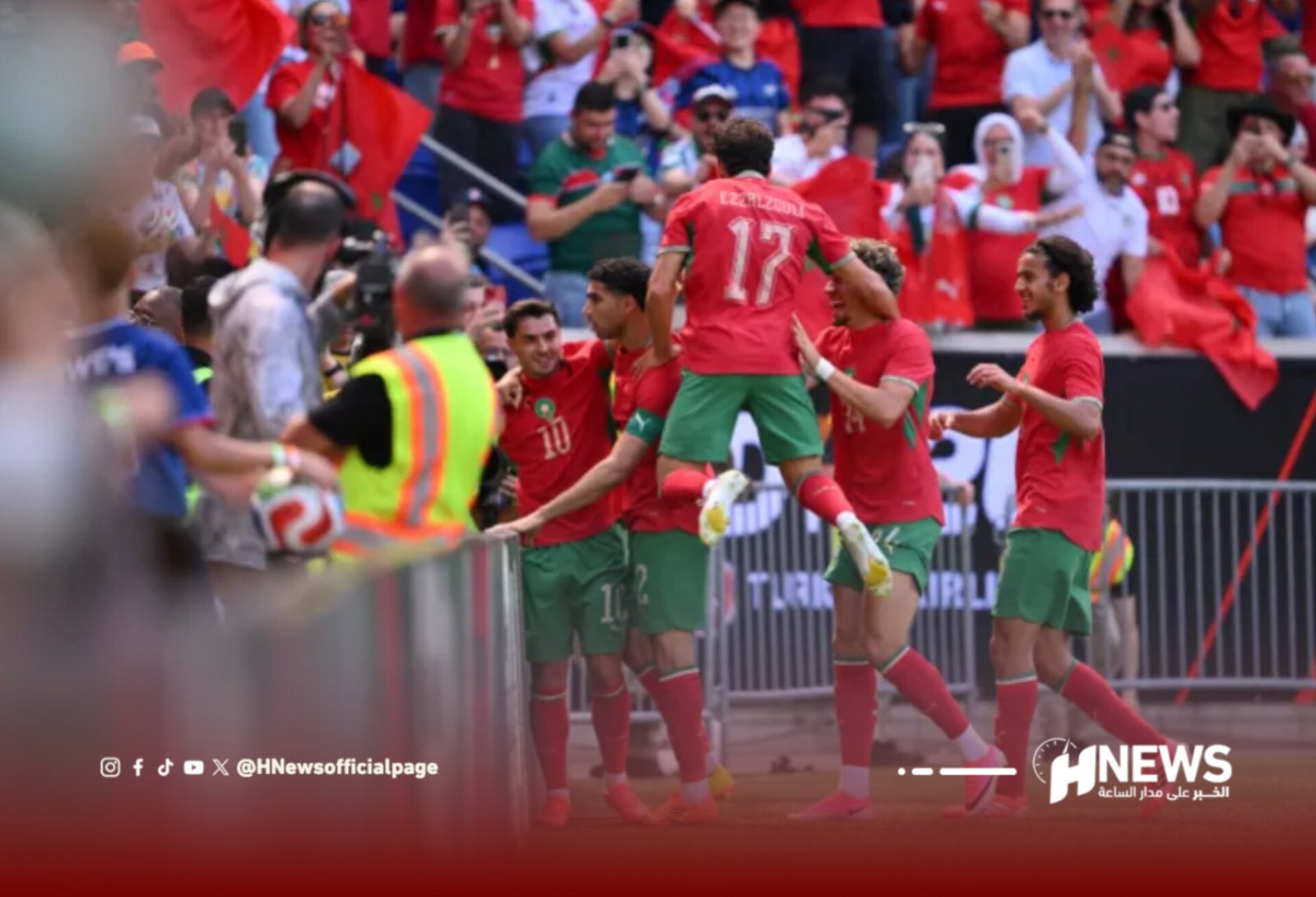 Soccer players in red uniforms celebrate a goal near the sideline as a cheering crowd watches from behind barriers.