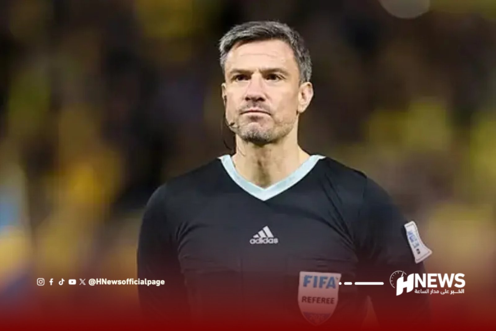 Male football referee in a black shirt with a FIFA badge on the chest, standing on the pitch during a match.