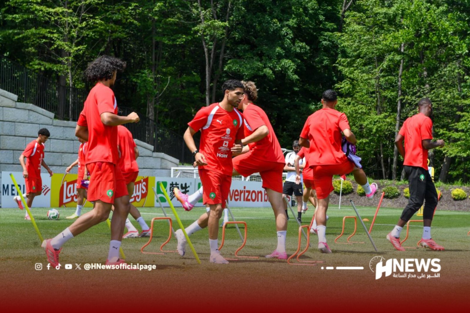 Soccer players in red uniforms doing agility drills with small hurdles on a grassy field during practice, with trees in the background and a news logo at the bottom right.