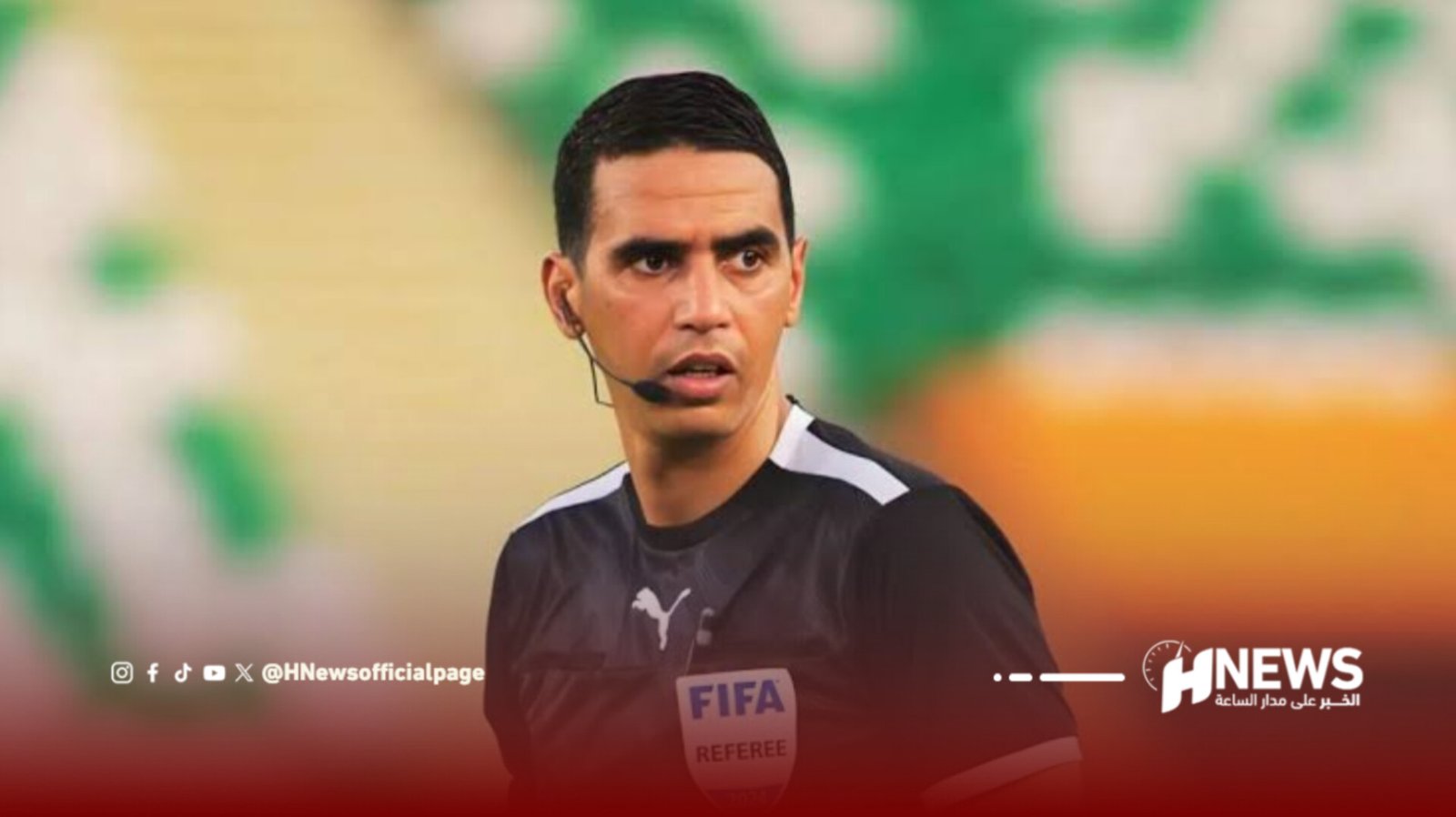 Football referee wearing a headset mic and FIFA badge on a blurred stadium background, looking to the left.
