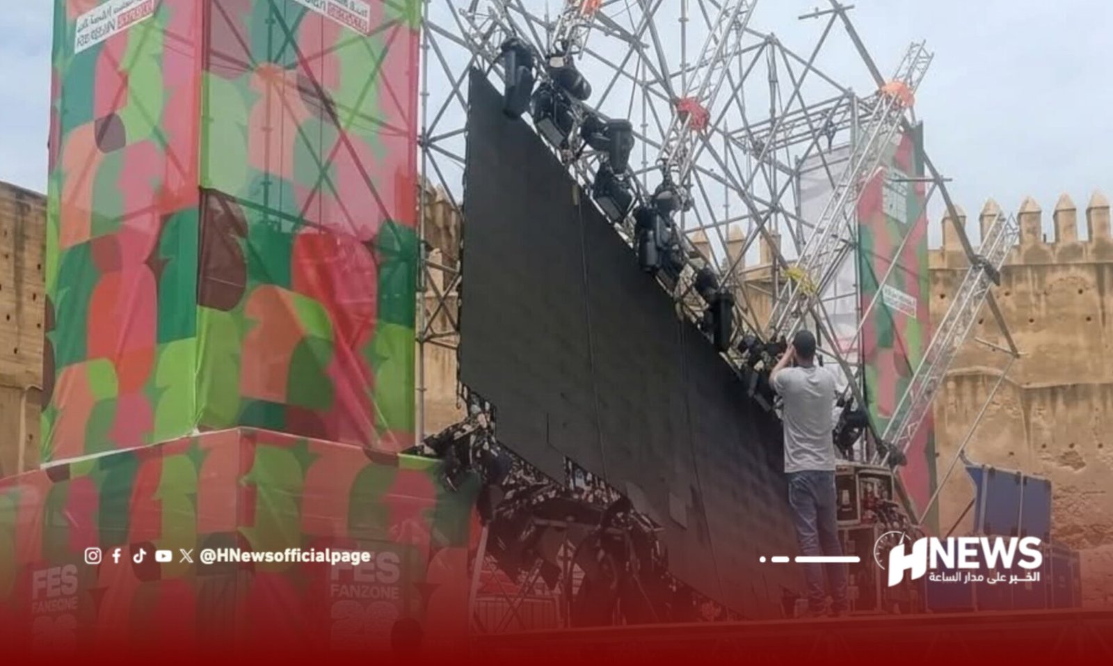Stage crew raising a large black backdrop on a metal scaffold at an outdoor event, with colorful banners on the sides.
