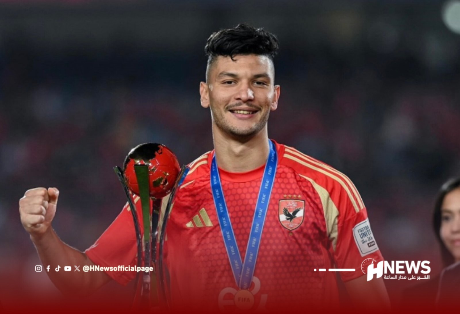 Footballer in red jersey holding a trophy and wearing a blue medal ribbon, celebrating victory on the field with a blurred crowd behind him at night