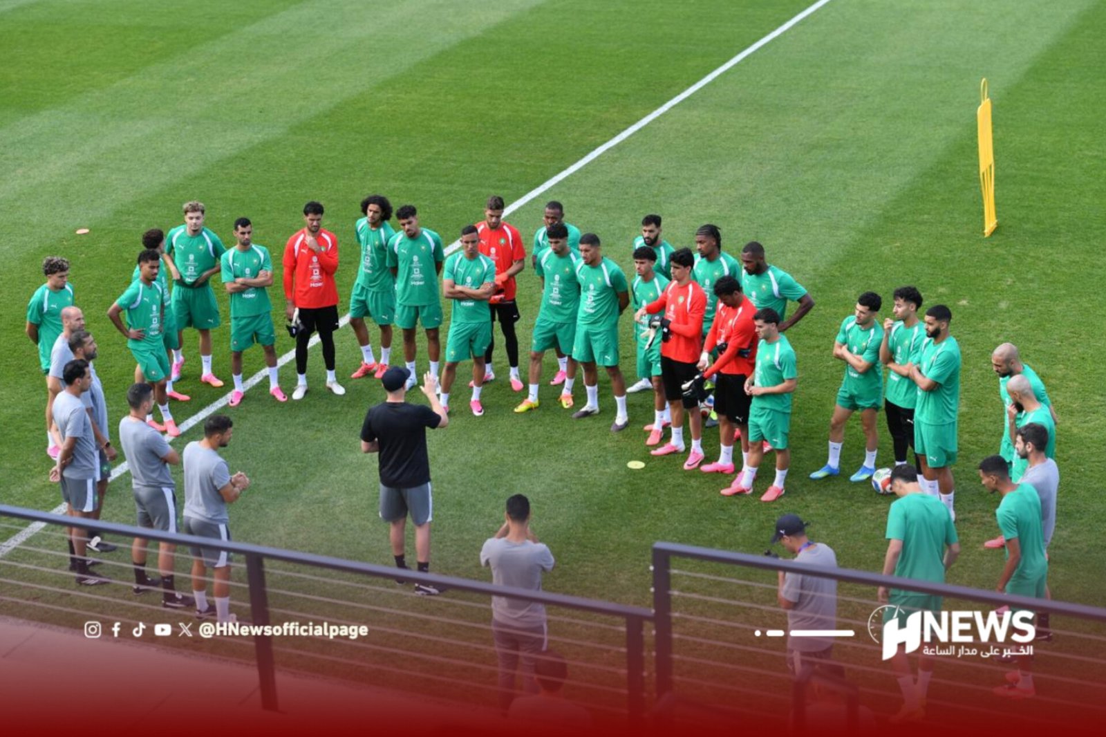 Soccer team in green uniforms lined up on a grass field for a briefing, with coaches at left and red-jersey goalkeepers among them.