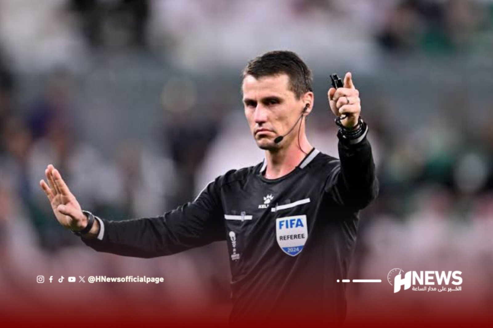 Football referee in a black kit with a FIFA badge raises both arms on the field, signaling a call, with a headset mic visible.