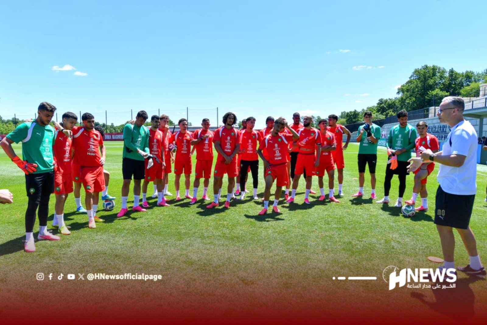 Soccer players in red uniforms form a line on a sunny training field while a coach speaks from the right.
