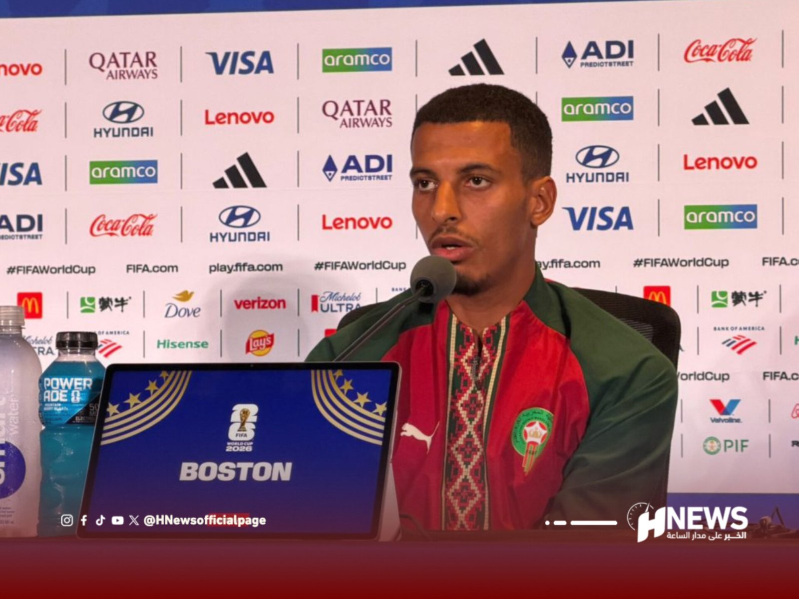 Male athlete in a red and green outfit speaks at a press conference with a sponsor backdrop behind him.