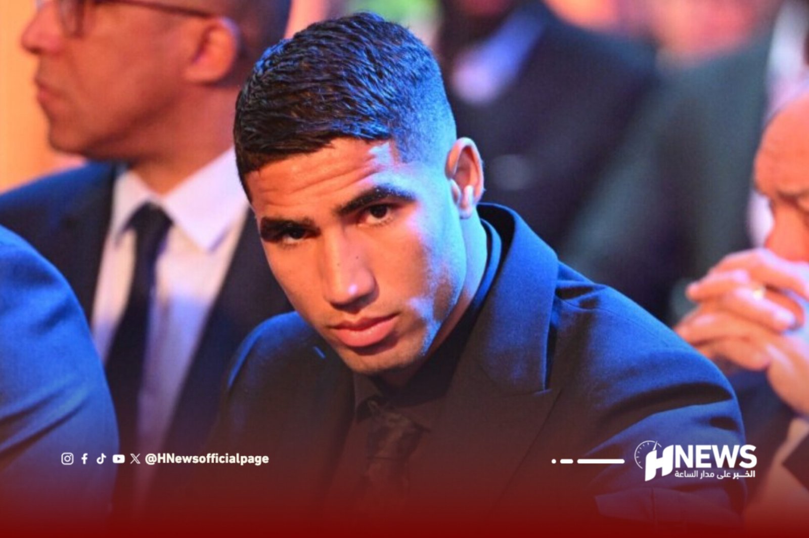Young man in a dark suit looking down at an event, blue stage lighting and HNews watermark in the corner.
