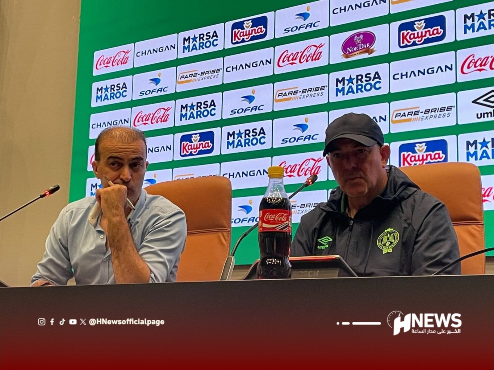 Two men sit at a press conference table against a green sponsor backdrop; one rests his chin on his hand, the other wears a cap, with a Coca‑Cola bottle on the table between them.