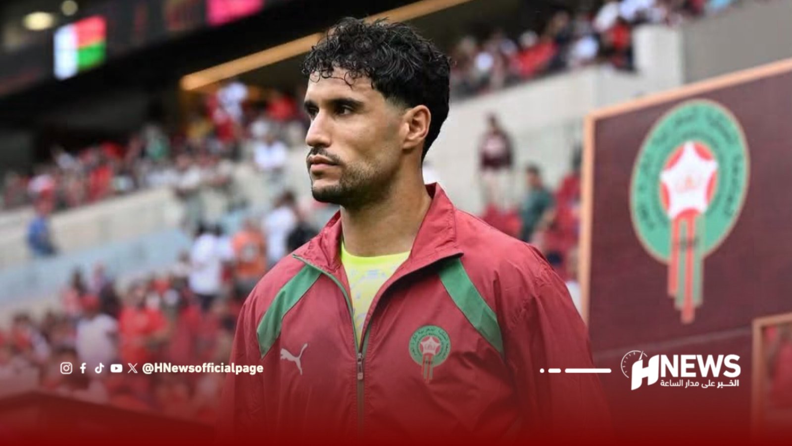 Athlete wearing a red jacket with green details walks on a pitch with a crowded stadium behind him.