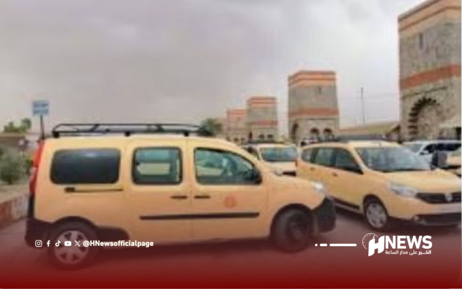 Row of yellow taxis parked in a lot with a large beige building featuring arches in the background.
