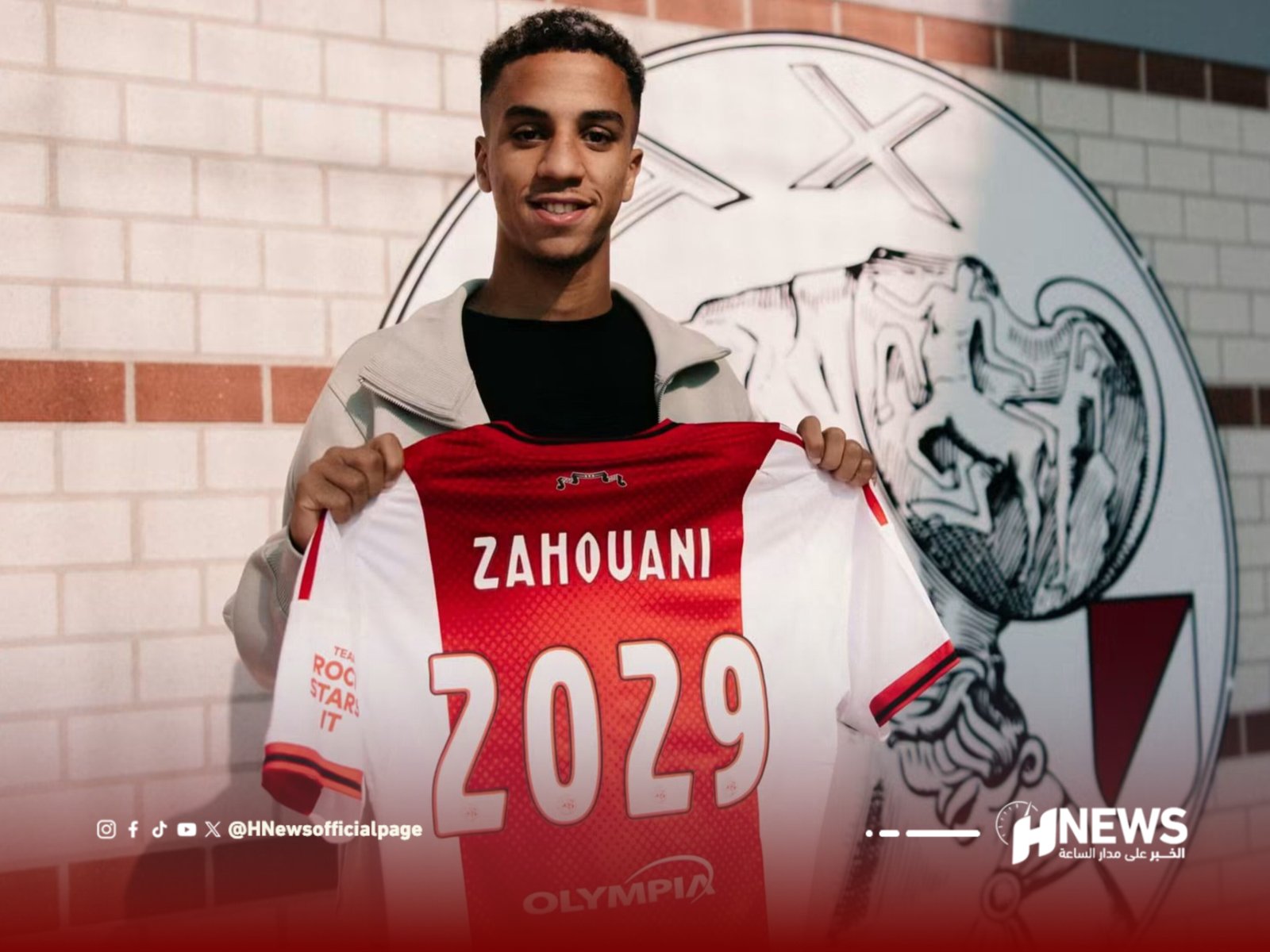 Young man posing and displaying a red and white sports jersey with the name 'ZAHOUANI' across the chest in front of a mural.